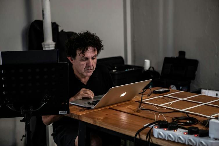 A curly-haired man sits concentrating as he works on a laptop in a studio.