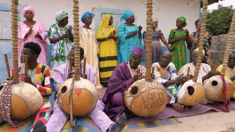 Six men in traditional west African robes and headgear walk in a group playing wooden stringed instruments in front of a rural homestead. Behind them stands a row of women in traditional attire, clapping.