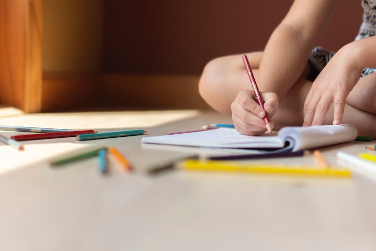 A child sits on the floor, with coloured pencils and a notebook.