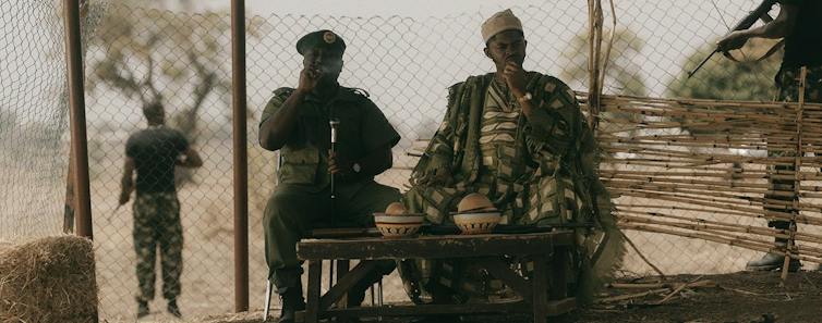 Two men sit on a porch at a small table with plates. One is in military uniform. An armed soldier keeps watch behind them.