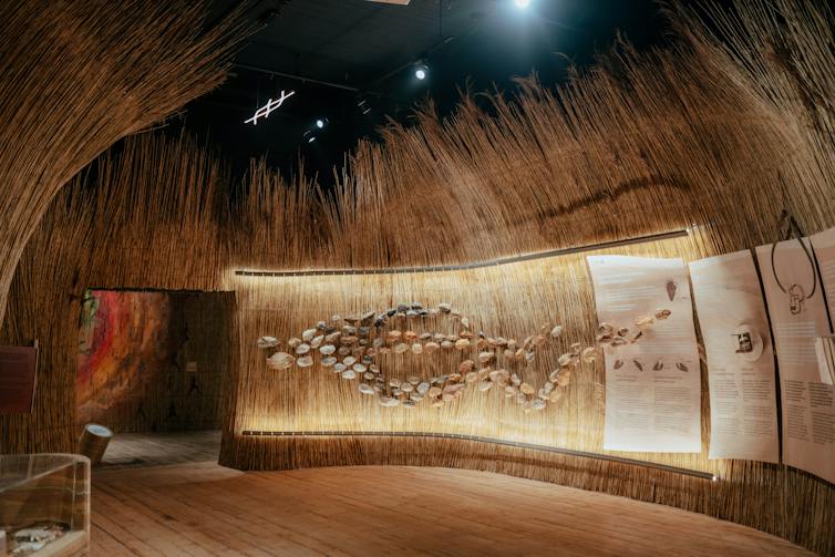 A fully thatched room that resembles the inside of a large hut. Pieces of stone have been affixed to one wall in a braided pattern and there are text boxes next to the design.