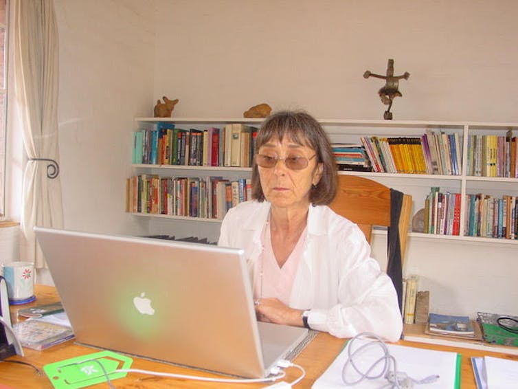 A woman sits at a desk, focused on reading from a computer screen in front of her, rows of books lining the walls behind her.